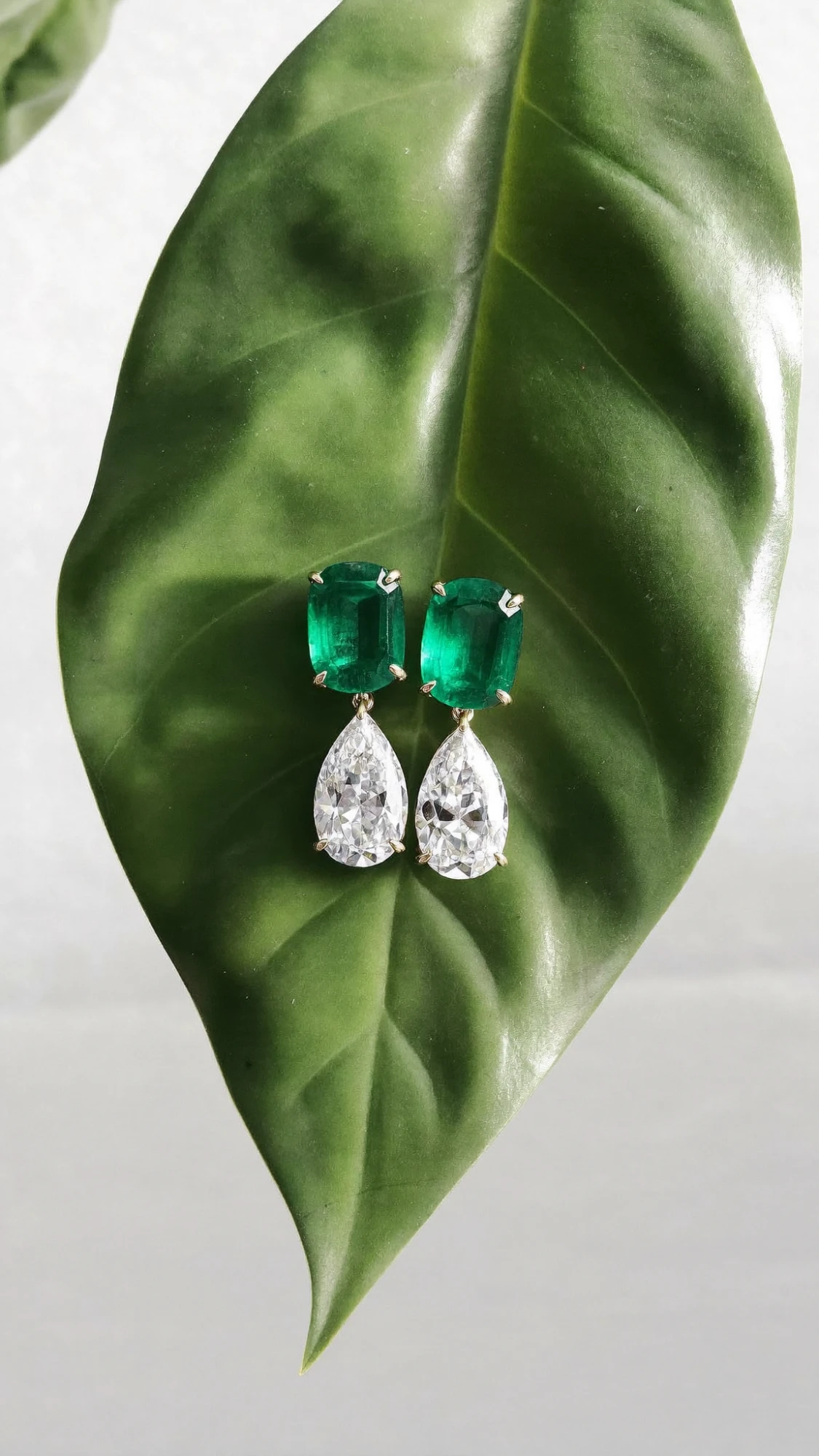 Emerald and pear-cut diamond drop earrings resting on a glossy green leaf
