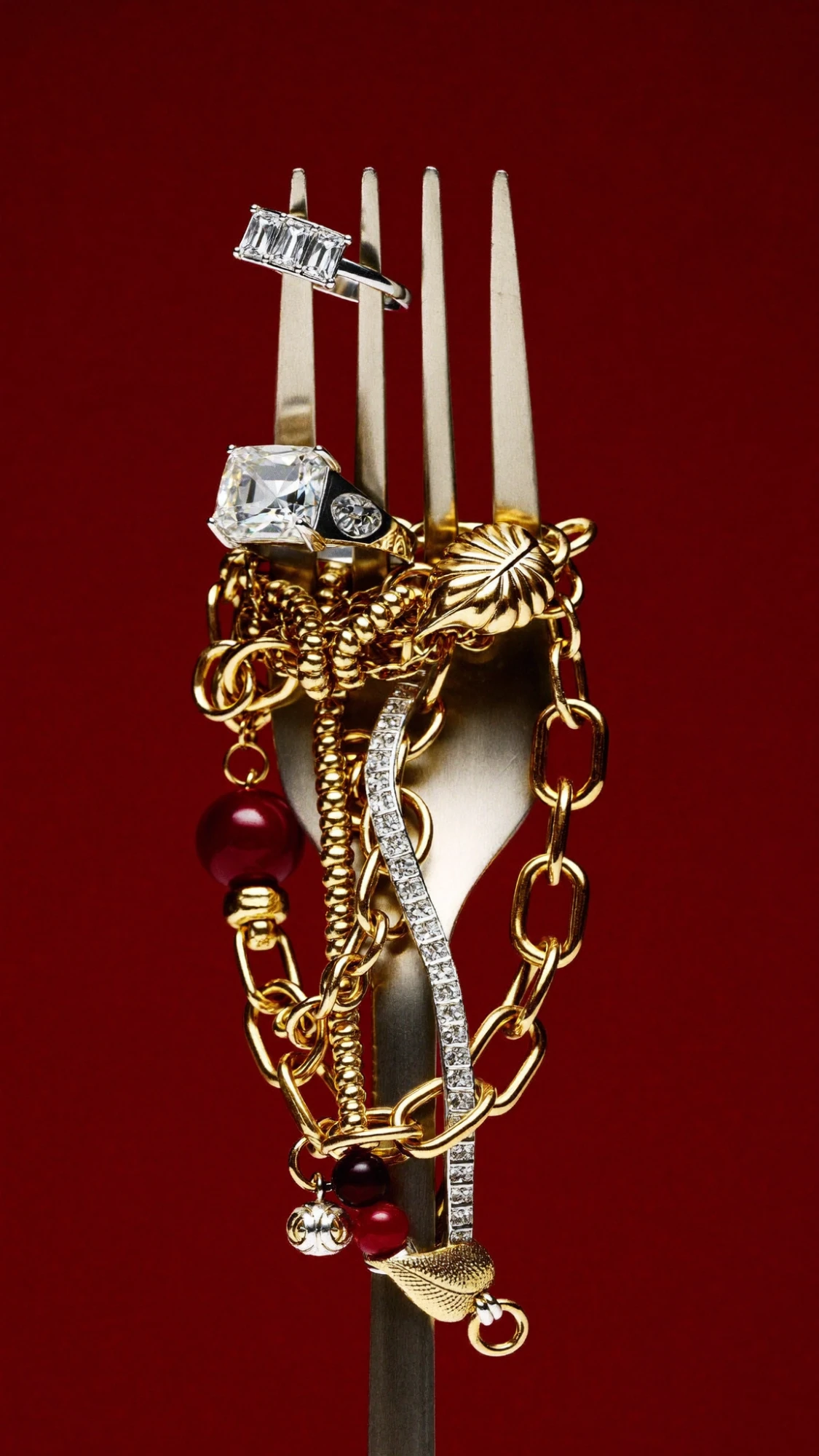 Gold chains, pearls and diamond rings wrapped around a silver fork against deep red